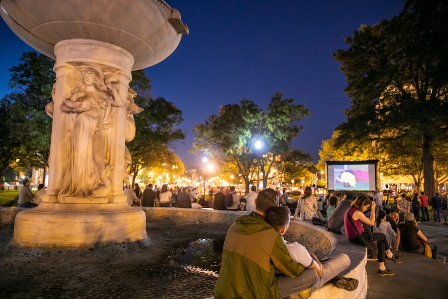 A couple sits on the edge of a fountain in Dupont Circle, part of an audience watching an outdoor movie. 