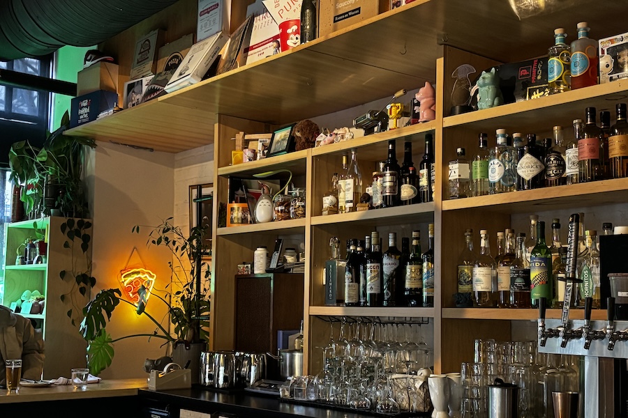 A cozy bar with wooden shelves stocked with liquor bottles, glassware and eclectic decorations, including a neon pizza sign.