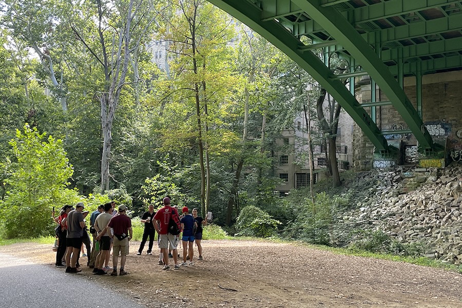 A guided group hike pauses under a green steel bridge in Rock Creek Park near stone ruins and graffiti-covered walls.