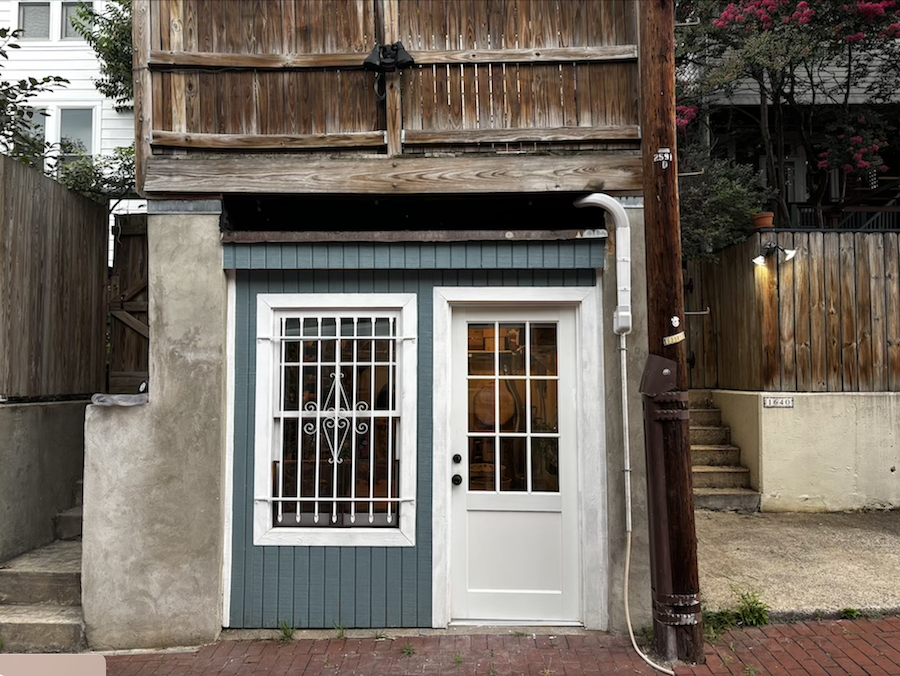 A small, rustic storefront with a white door and barred window sits along a brick alley, surrounded by wood fencing and stairs.