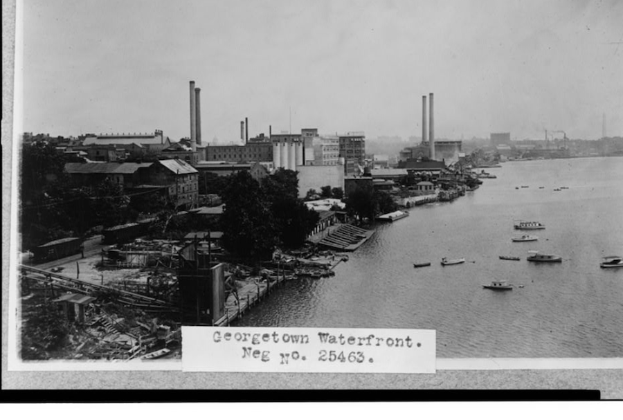lack-and-white photo of the historic Georgetown waterfront with industrial buildings, smokestacks and boats on the Potomac River.