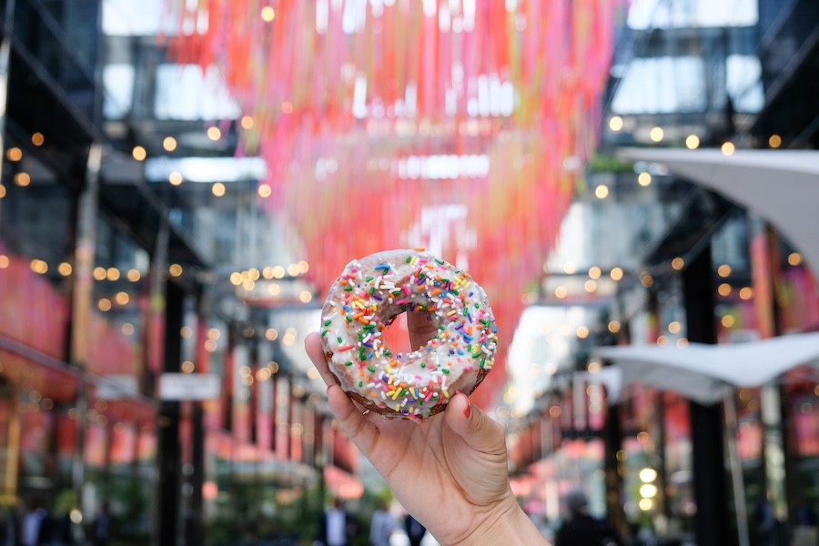 A hand holds up a sprinkle-covered donut in front of colorful streamers hanging above Palmer Alley in CityCenterDC.