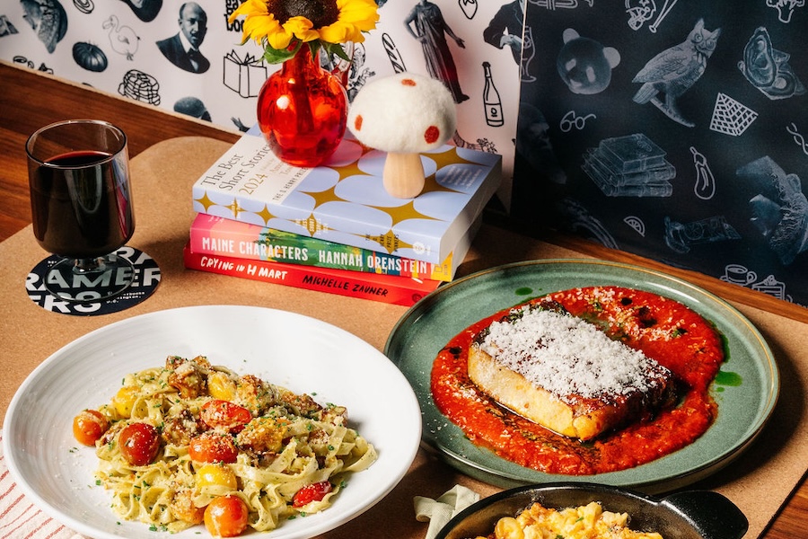 A table at Kramers Café features plates of pasta, a glass of wine and a stack of books beside a small vase of flowers.