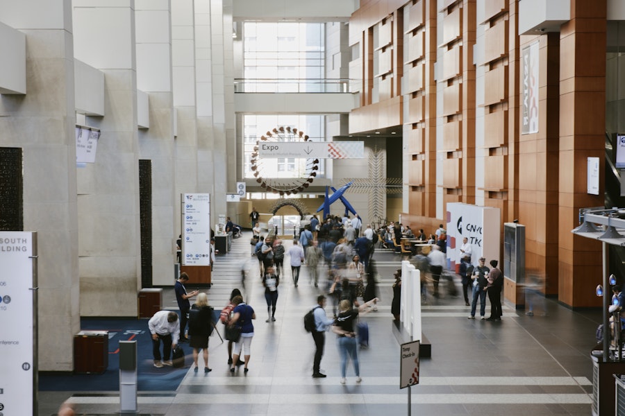 A busy convention center atrium with people walking, talking, and checking in near large signage and modern architectural features.