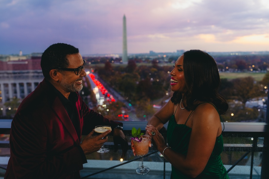 A couple enjoys cocktails on a rooftop terrace with the Washington Monument visible in the background at dusk.