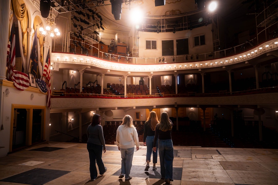 People stand on stage at a historic theater, gazing up at the balconies, the spotlights and the velvet drapery.