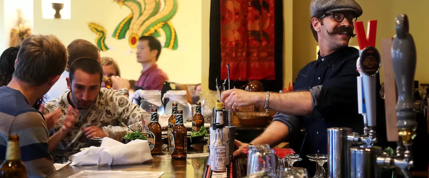 Bartender with a mustache serves drinks to patrons at a busy bar, decorated with vibrant artwork in the background.