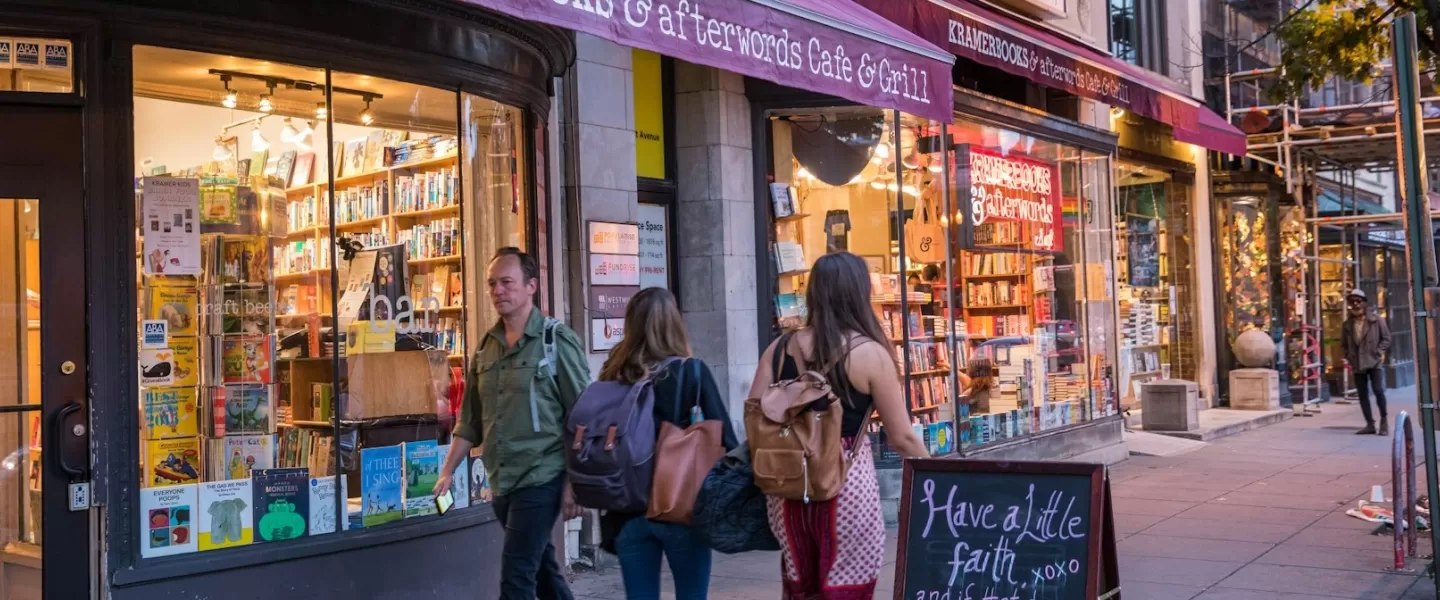 People stroll past the inviting storefront of Kramerbooks & Afterwords Cafe in Dupont Circle, with shelves of books visible through large windows and a chalkboard sign on the sidewalk.