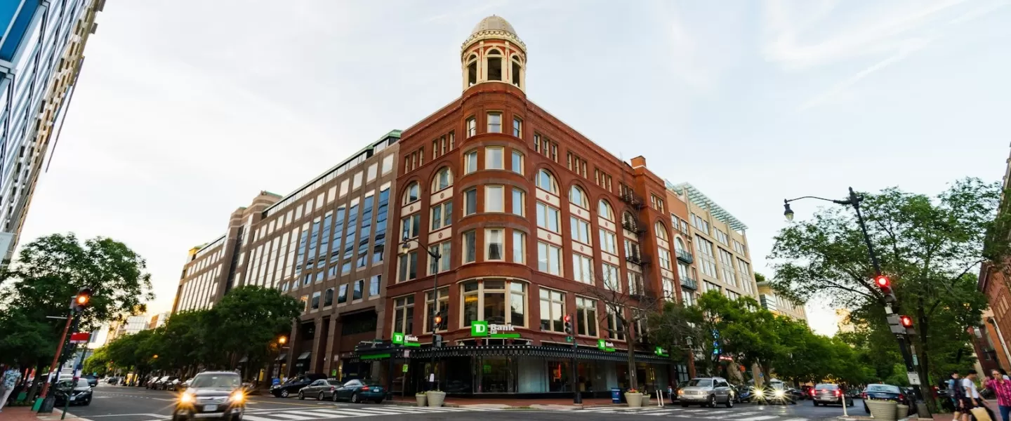 A striking red-brick building with arched windows and a corner turret stands at a busy downtown Washington, DC intersection, surrounded by modern glass structures.