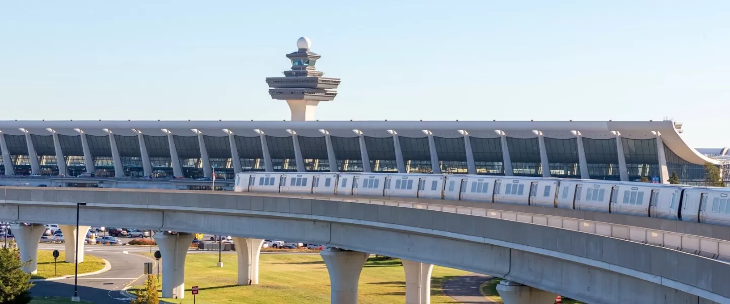 Silver Line Metro train traveling on elevated tracks with Washington Dulles International Airport terminal and air traffic control tower in the background.