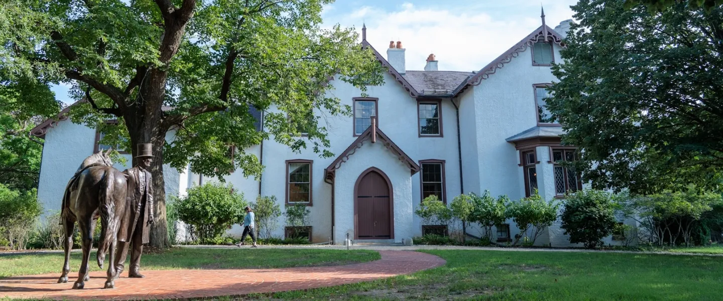 A statue of Abraham Lincoln with his horse stands before the historic Lincoln’s Cottage, surrounded by trees and greenery.