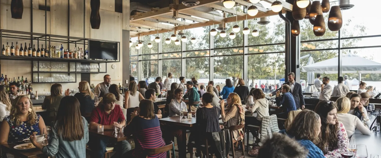 Interior scene of people dining and drinking at District Winery, with the river in the background. 