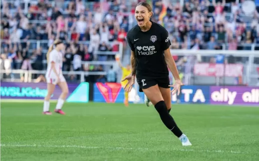 A Washington Spirit player celebrates a goal with an intense expression as the crowd cheers in the background.
