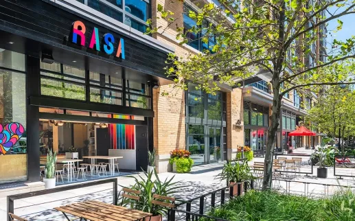 The exterior of Rasa restaurant in Mount Vernon Square features bright modern decor and a sunny outdoor patio with seating and greenery.
