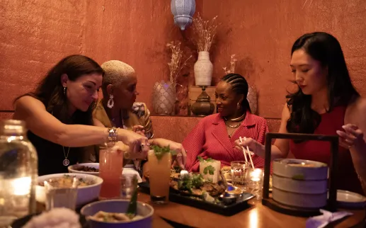 A group of friends share small plates and colorful cocktails at Doi Moi in Washington, DC, seated around a candlelit table with warm, textured walls in the background.