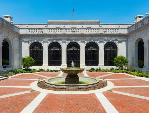 A round fountain sits at the center of a bright brick courtyard surrounded by white stone arches and large windows in Washington, DC.