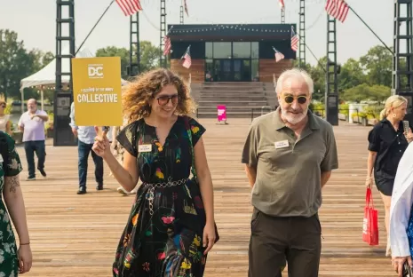 A group strolls along a sunny boardwalk, led by a smiling participant holding a "Collective" sign, under festive string lights and American flags.