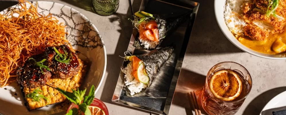 An overhead view of a diverse spread of Japanese dishes, featuring sushi rolls, rice bowls, and various appetizers, along with vibrant cocktails and meticulously plated dishes on a modern table setting.