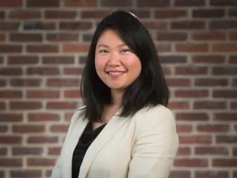 A person with straight dark hair wearing a cream blazer, smiling in front of a brick wall.